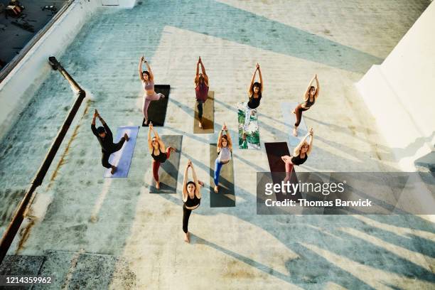 overhead view of yoga class in warrior pose while practicing on rooftop - personen gruppe von oben stock-fotos und bilder