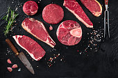 Various cuts of meat, shot from the top on a black background with salt, pepper, rosemary and knives, with copy space