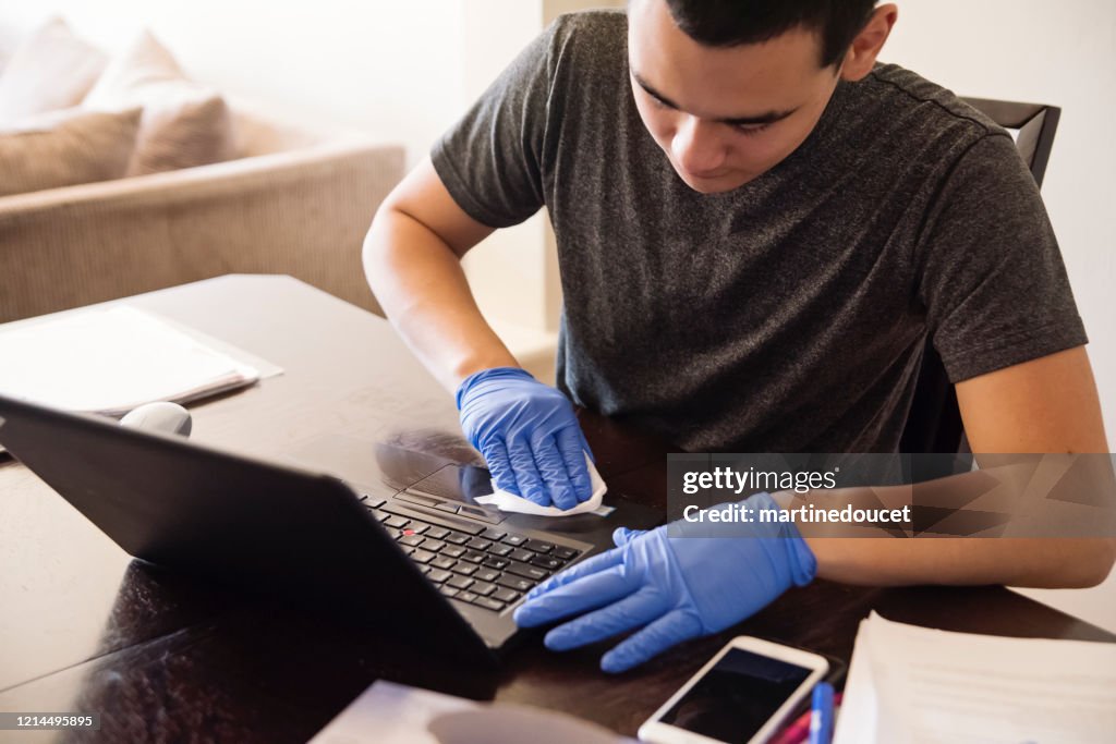 Teenage boy with gloves wiping laptop while studying from home.