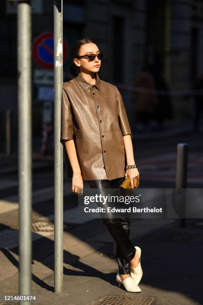 Beatrice Gutu wears sunglasses, a bracelet, a brown leather shirt, black leather pants, a caramel-brown crocodile pattern clutch, white pointy pumps...