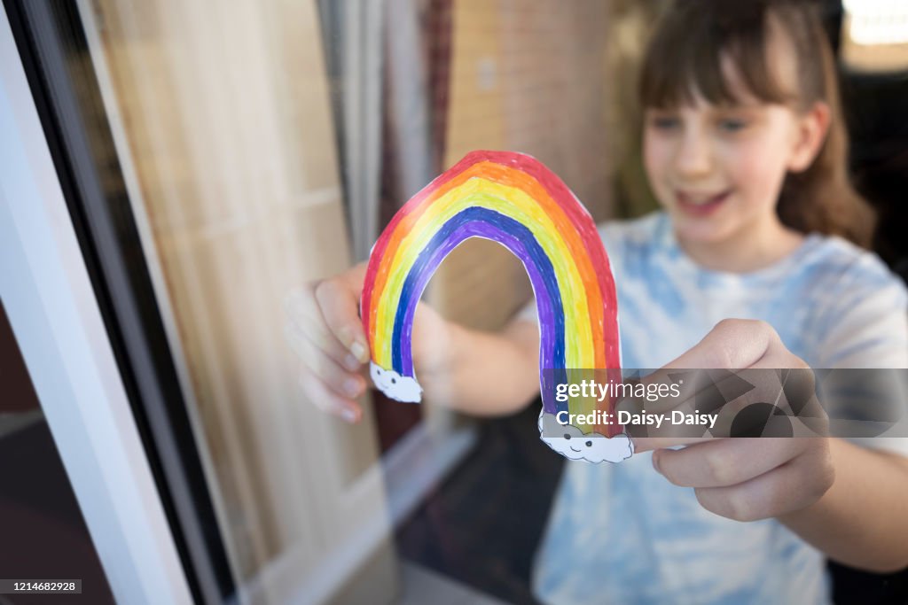 Girl Putting Picture Of Rainbow In Window At Home During Coronavirus Pandemic