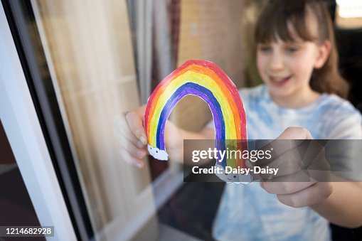 Girl Putting Picture Of Rainbow In Window At Home During Coronavirus Pandemic Girl Putting Picture Of Rainbow In Window At Home During Coronavirus Pandemic