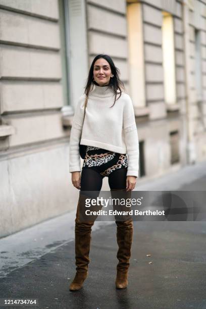 Guest wears a white turtleneck wool pullover, a bag, brown suede thigh high boots, leggings with leopard print, earrings, outside Alessandra Rich,...
