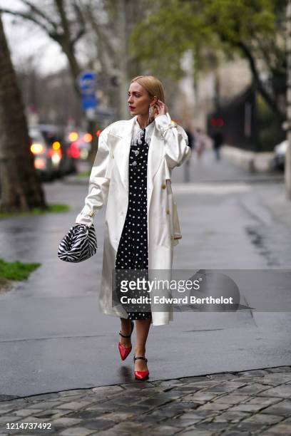 Viktoria Rader wears long bejeweled earrings, a white long coat, a black and white zebra print puff bag, a black and white dress with polka dots, red...