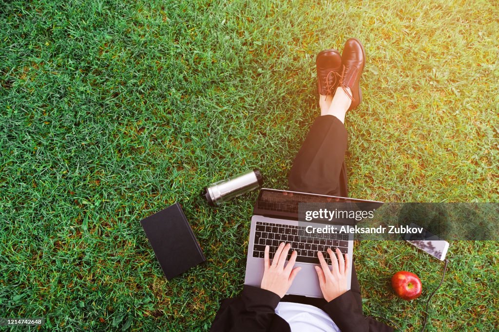 A girl in the Park sitting on the grass and working on a laptop, a woman's hands on the keyboard. Near mobile phone, Apple, notebook. Student studying outdoors. Distance learning concept