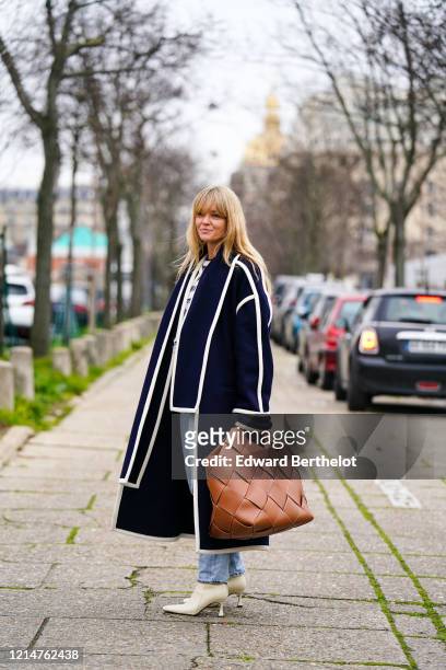 Jeanette Madsen wears a dark blue long coat, a brown leather woven large bag, blue denim jeans pants, white pointy shoes, outside Loewe, during Paris...