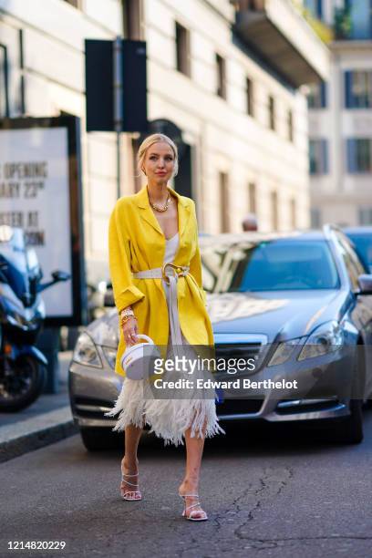 Leonie Hanne wears earrings, rings, bracelets, sunglasses, a chain necklace, a white dress with a wide neckline and decorated with white feathers, a...