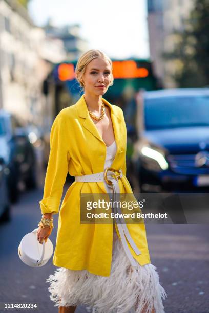 Leonie Hanne wears earrings, rings, bracelets, sunglasses, a chain necklace, a white dress with a wide neckline and decorated with white feathers, a...