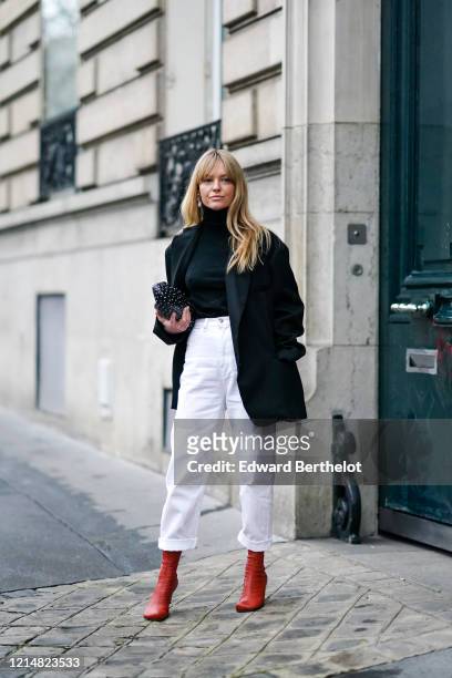 Jeanette Madsen wears earrings, a black turtleneck pullover, a black oversized blazer jacket, white pants, red leather boots, a black bag with polka...