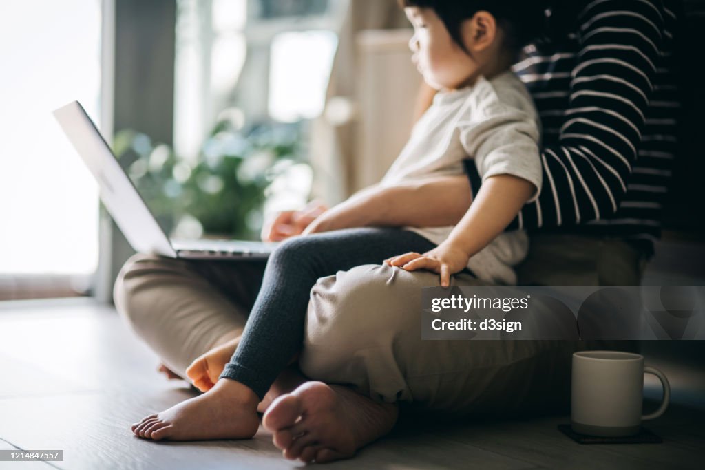 Cropped shot of a young Asian mother using laptop and working from home while taking care of little daughter in self isolation during the Covid-19 health crisis