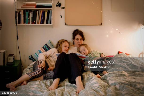 full length of mother reading picture book while sitting with children in bedroom - reading stock-fotos und bilder
