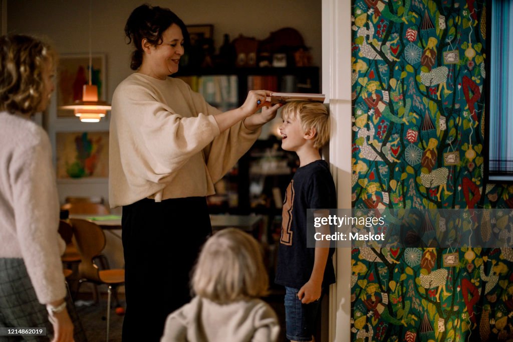Mother measuring height of smiling son with book on wall