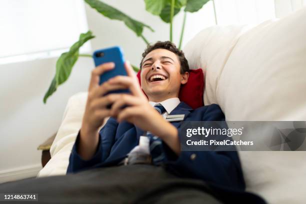 adolescente niño de la escuela en uniforme acostado en el sofá riendo usando el teléfono móvil - meme fotografías e imágenes de stock