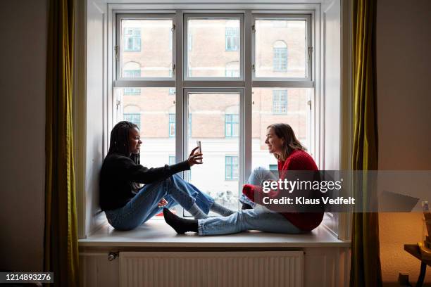 young female roommates using smart phones at apartment window - common aims stock-fotos und bilder