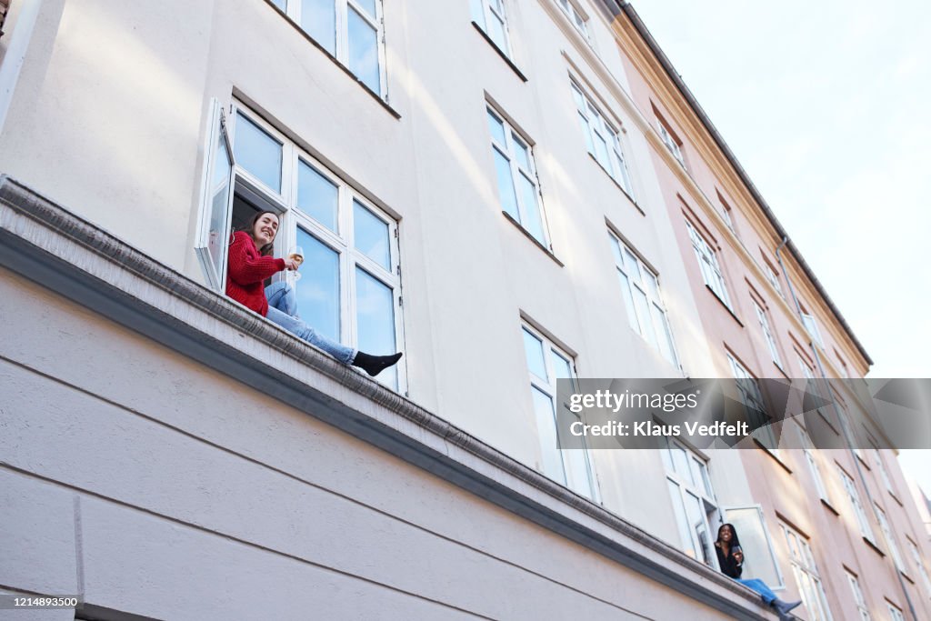 Portrait young female neighbors drinking wine and talking from apartment windows
