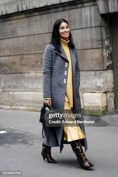 Caroline Issa wears a yellow wool turtleneck woven pullover, a gray long coat, a black leather bag, a yellow lustrous silky skirt, black and yellow...