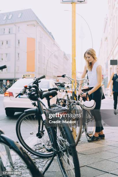 full length of young entrepreneur locking bicycle on sidewalk in city - fahrradschloss stock-fotos und bilder