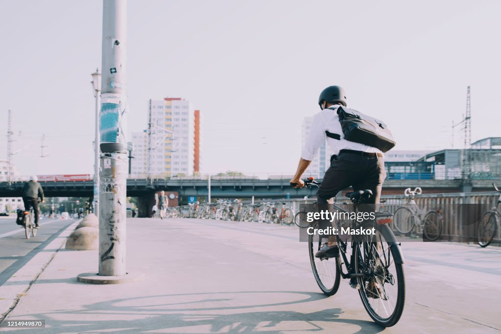 Rear view of businessman riding bicycle on sidewalk against clear sky in city
