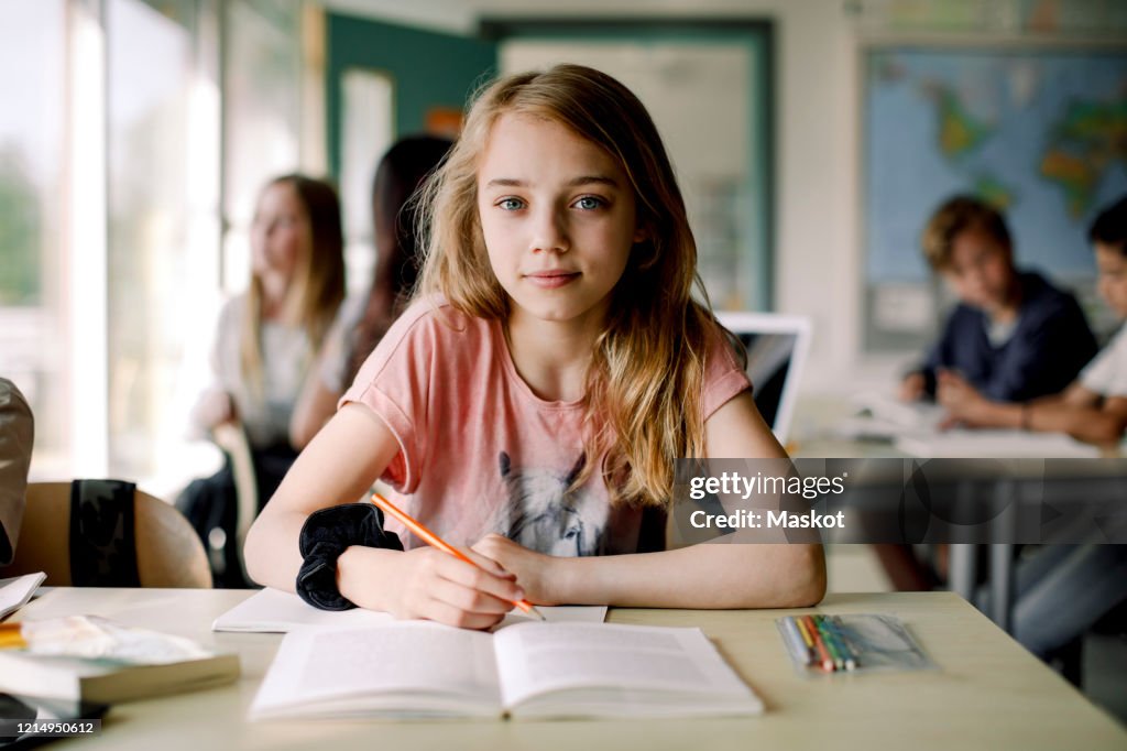 Portrait of female student writing in book while sitting at table in classroom