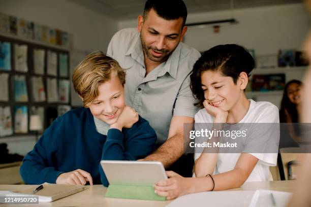 smiling male tutor with students using digital tablet while sitting in classroom - middle school stock-fotos und bilder