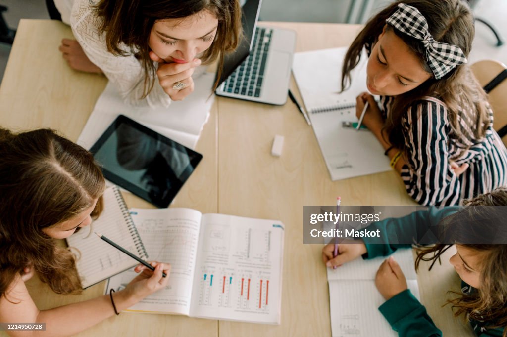 High angle view of students with female tutor studying from book at table in classroom