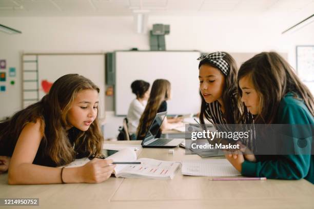 female students studying from book while sitting by table in classroom - junior high student stock pictures, royalty-free photos & images