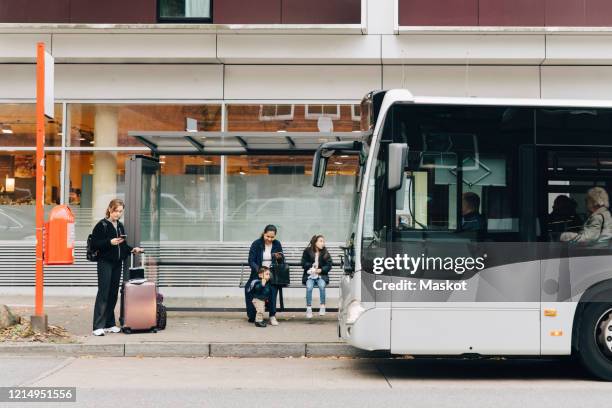 full length of woman with luggage and family waiting for bus at bus stop in city - fermata di autobus foto e immagini stock