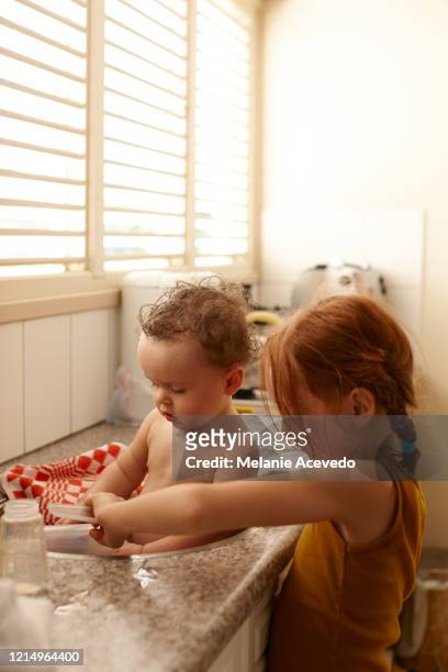 Baby playing with younger red headed sister in sink looking down at water
