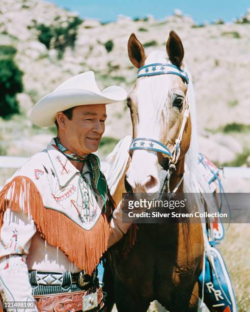 Roy Rogers , US actor and singer, wearing a white cowboy hat, red neckerchief and fringed western shirt, posing beside his palomino horse, Trigger ,...