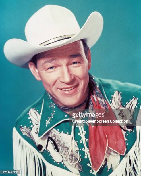 Roy Rogers , US actor and singer, wearing a white cowboy hat, red neckerchief and fringed western shirt in a studio portrait, against a blue...