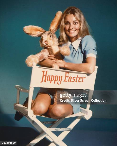 Sharon Tate , US actress, wearing a blue dress, with a white collar, holding a toy rabbit and sitting in a director's chair with the legend, 'Happy...