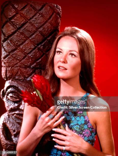 Shelley Fabares, US actress, wearing a floral print sleeveless dress, holding a red flowers, posing beside a Polynesian sculpture in a studio...