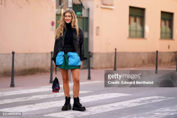 Emili Sindlev wears a black jacket, a black turtleneck pullover, green skirt with large blue pockets, a bag with red printed flames, black leather...