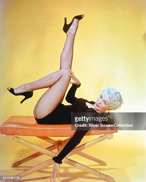 Sheree North , US actress, singer and dancer, wearing a black long-sleeved leotard with fishnet stockings, laying on her back on a stool, with her...