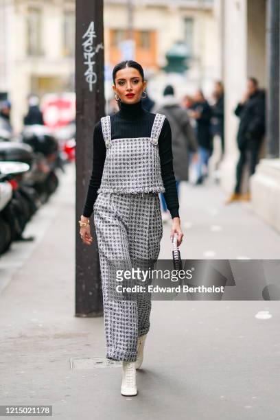 Nathalie Fanj wears earrings, a black turtleneck pullover, a silver knitted top with squared patterns, gray pants, a bejeweled bag, white shoes,...