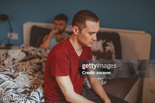 Disappointed man sitting on bed side, his boyfriend in bed Disappointed man sitting on bed side, his boyfriend in bed