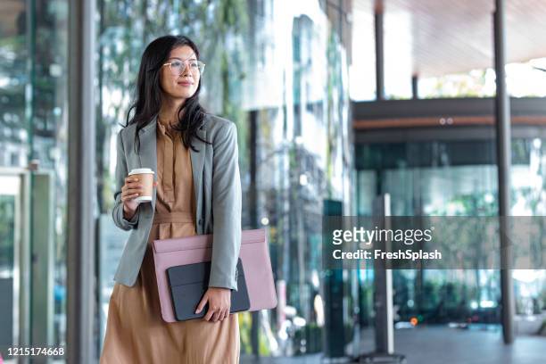 happy successful asian businesswoman holding a takeaway coffee cup and files on the street next to a glass building - professional occupation stock pictures, royalty-free photos & images