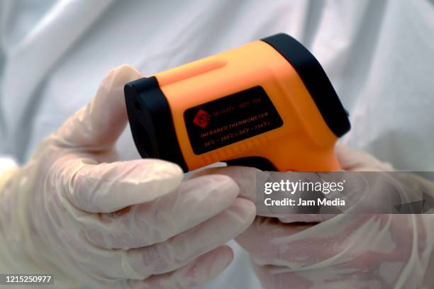 Volunteer medical worker dressed in a protective suit, mask, gloves and goggles holds a digital thermometer in a tent set up to test people for...