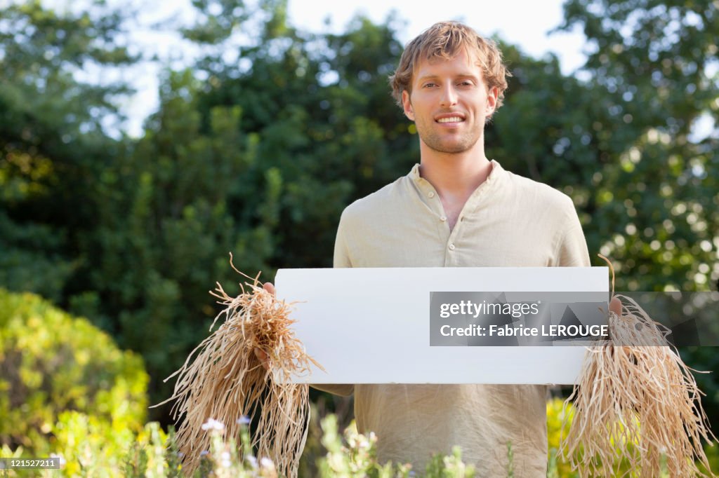Young man in scarecrow dressing and holding a blank placard in a field