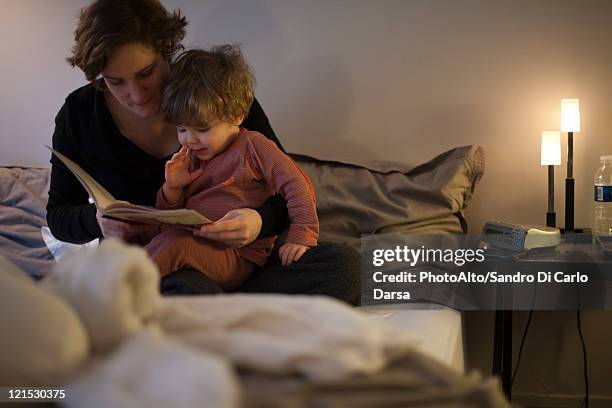 mother holding toddler son on lap, reading bedtime story in bed - kinderbuch stock-fotos und bilder