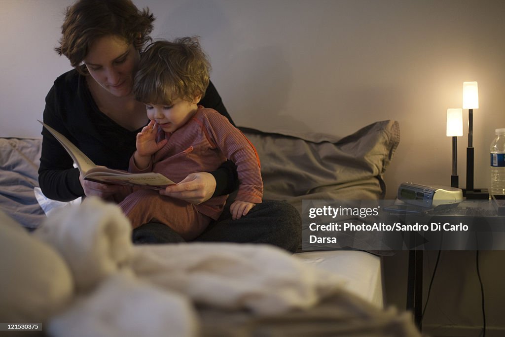 Mother holding toddler son on lap, reading bedtime story in bed