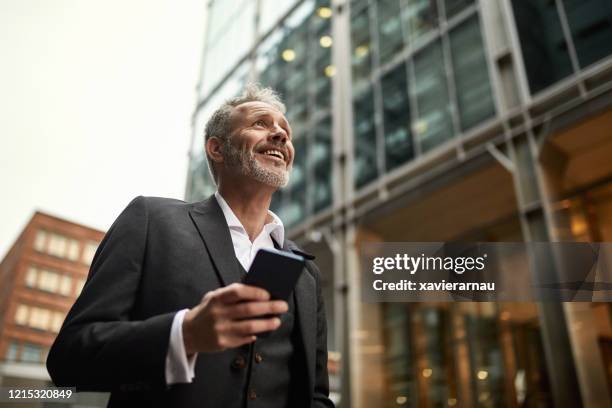 mature businessman smiling while using smart phone in london - man walking low angle stock pictures, royalty-free photos & images