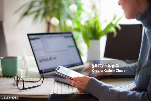 woman working from home using laptop computer while reading text message on mobile phone - computer stock-fotos und bilder