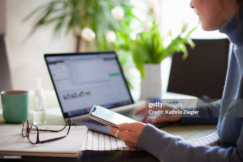Woman working from home using laptop computer while reading text message on mobile phone