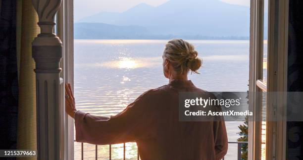 reife frau geht auf veranda über einen see bei sonnenaufgang - balcony view stock-fotos und bilder