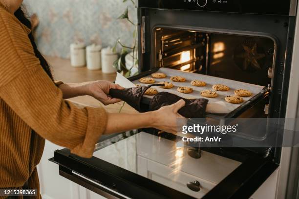 jovem mãe colocando uma bandeja cheia de biscoitos no forno - oven - fotografias e filmes do acervo