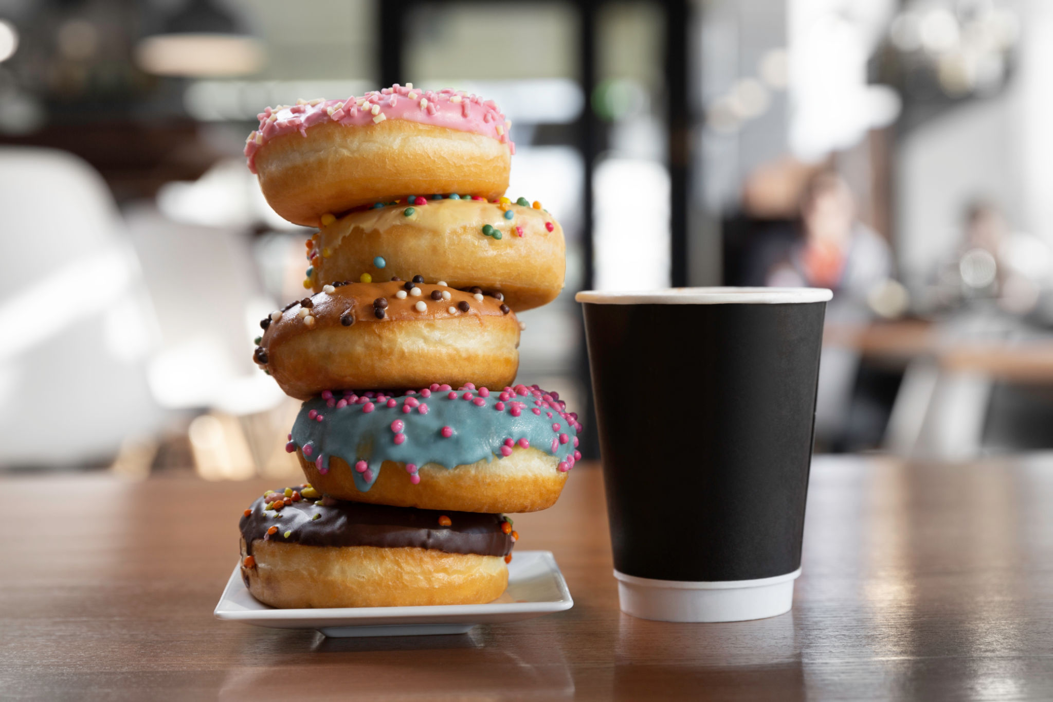 Colorful glazed donuts pyramid on a white plate standing close to black desposable paper coffee cup Colorful glazed donuts pyramid on a white plate standing close to black desposable paper coffee cup