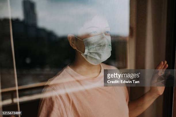 une femelle chinoise asiatique regardant à travers sa fenêtre de balcon avec son masque facial pendant l’ordre restreint de mouvement en malaisie - ordre de rester à la maison photos et images de collection