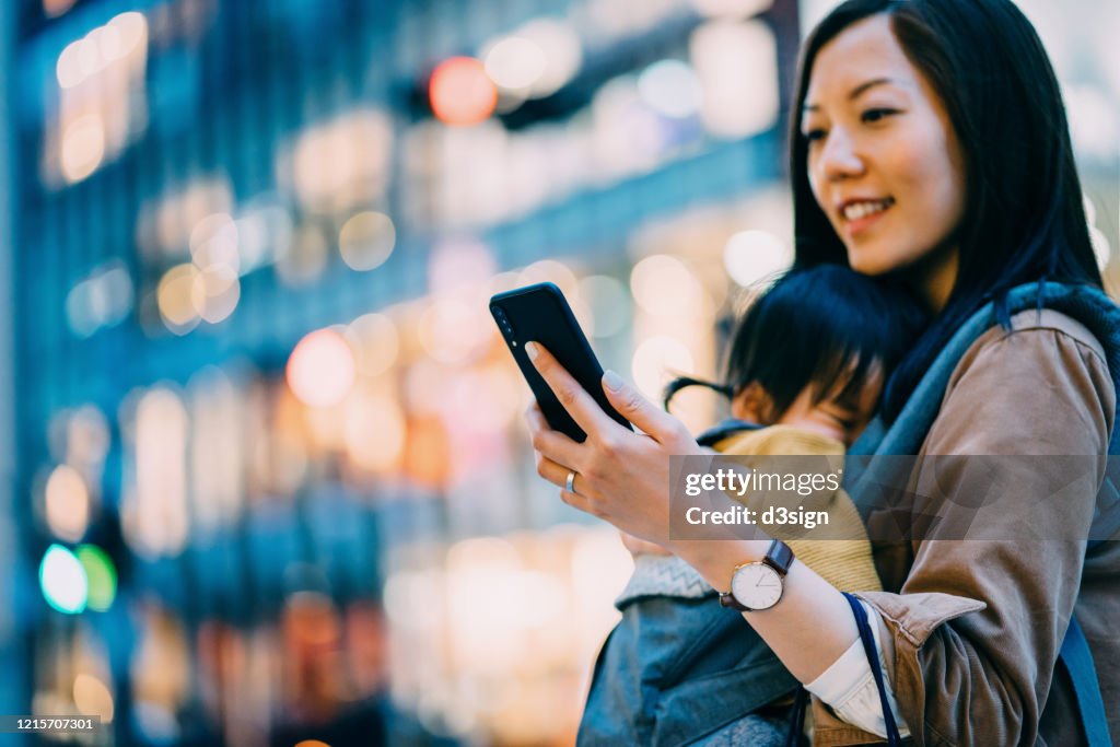Young Asian mother using smartphone in downtown city street while shopping with little daughter