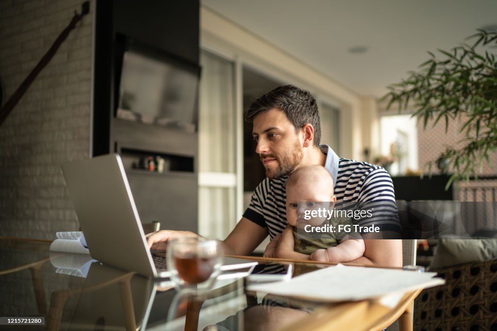 Mann hält sein neugeborenes Baby Sohn und arbeitet mit Laptop zu Hause